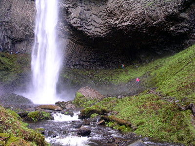 [A total whiteout of a huge stream of water hitting the ground with spray around it. There is then a gentle slope down which the water flows. A person wearing a red jacket stands in the vegetation on the right side, but none of the features are distiguishable since the person is small even in this closer view.]